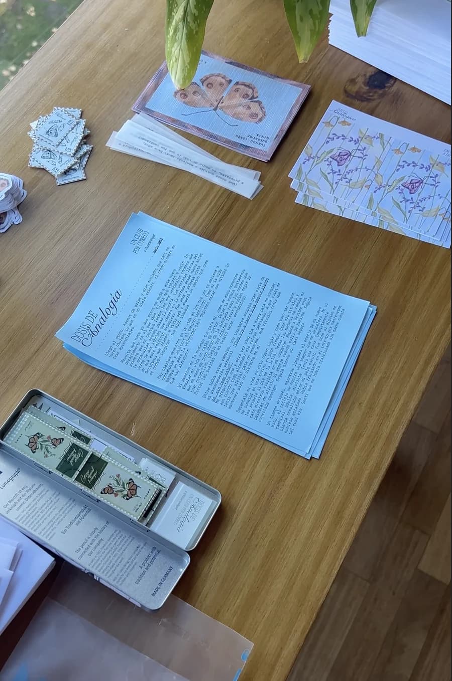 La mesa de trabajo con las piezas de la edición: la postal de la mariposa, una pila de estampillas, stickers recortados, las cartas impresas en celeste y una lata de lápices.